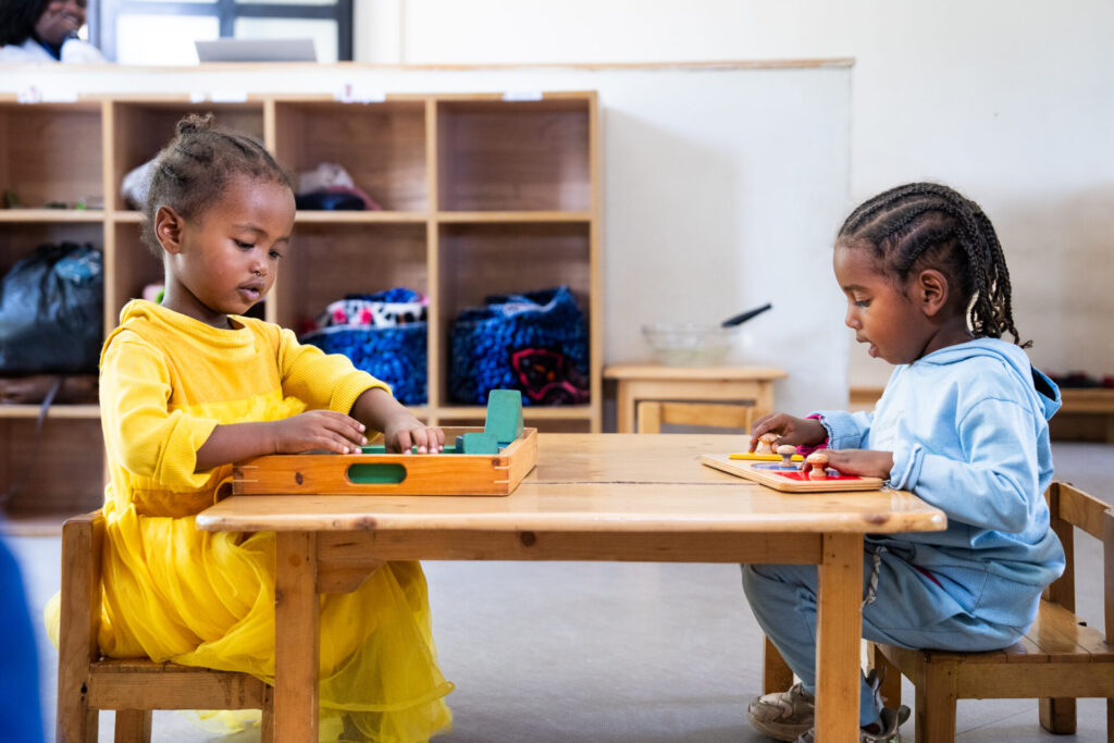 Two girls around 4 years old are sitting and writing in a kindergarten classroom.